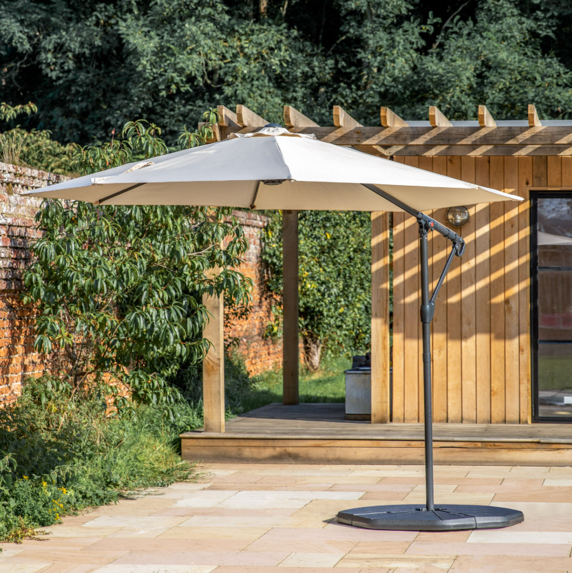 Beige patio umbrella on a stand in a garden setting with a wooden pergola and greenery.