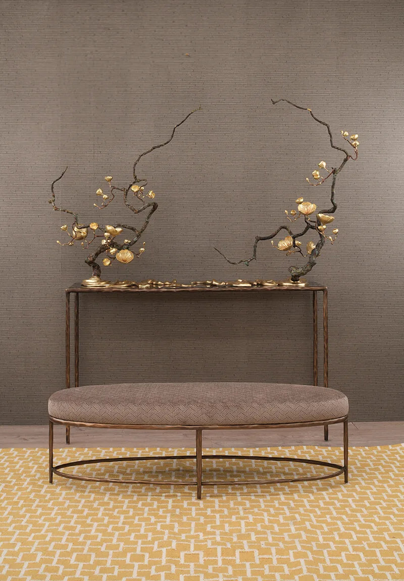 A modern console table with a round upholstered bench in front, set against a textured gray wall and a yellow patterned rug. The table and bench feature dark bronze metal frames with decorative gold-toned branches and blossoms mounted above the console.