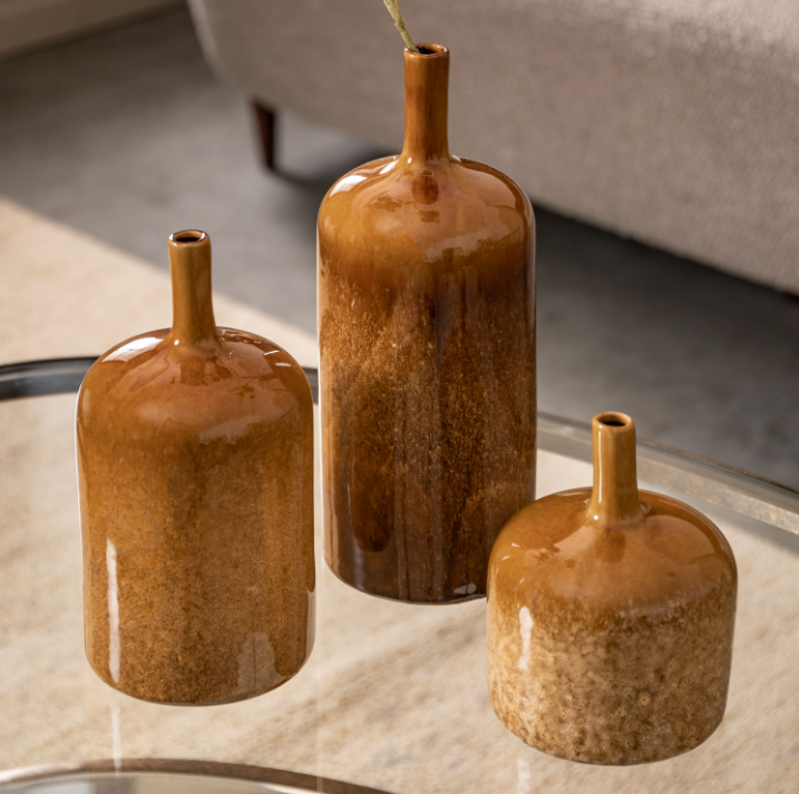 Three glossy ceramic vase-like bottles with short necks in different sizes, arranged on a glass table in a modern interior setting.