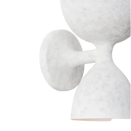 A close-up of a sculptural wall sconce with a textured, stone-like finish in off-white and light gray, featuring a round wall plate and a flared dome shade.